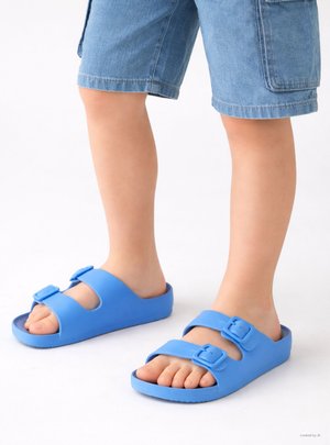 Bambino che indossa sandali blu regolabili con doppie cinghie e pantaloncini, in piedi su una superficie bianca.
