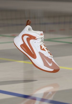 Hvid og orange Nike basketballsko, der svæver over en poleret indendørs bane med farverige afgrænsningslinjer.