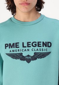 Tirkizni dukserica s okruglim izrezom i vezom "PME LEGEND AMERICAN CLASSIC" s grafikom krila u tamnoplavoj boji.