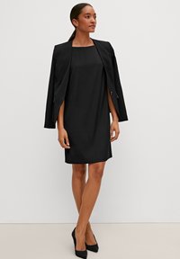 Schwarz Kleid mit quadratischem Ausschnitt, kurzen Ärmeln, aus glattem Stoff. Getragen mit einem passenden schwarzen Blazer und schwarzen hohen Schuhen.