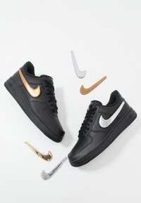 Baskets Nike Air Force 1 noires avec logos Nike Swoosh métalliques interchangeables en or et argent sur fond blanc.