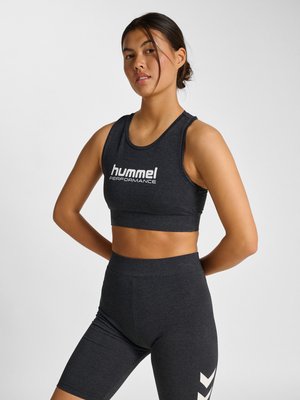 Kobieta w ciemnoszarym sportowym krótkim topie z napisem "hummel PERFORMANCE" i dopasowanych szortach z wysokim stanem, stojąca z rękami za plecami.