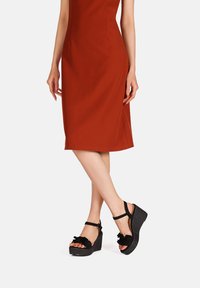 Sandali con zeppa nera e piattaforma texturizzata, con accenti floreali e cinturino alla caviglia. Indossati con un vestito aderente color ruggine, con una fessura laterale.