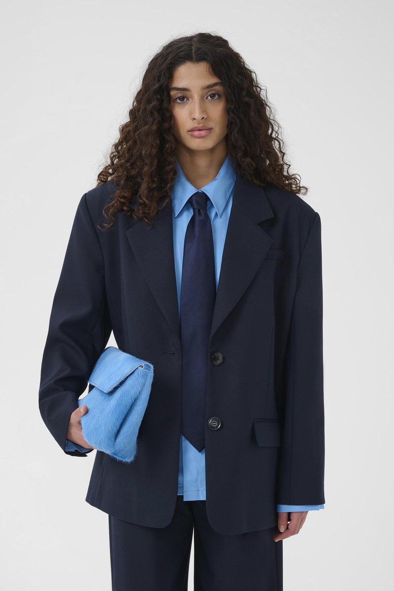 Γυναίκα με σγουρά μαλλιά που φορεί υπερμεγέθη ναυτικό κοστούμι, φωτεινό μπλε πουκάμισο, ναυτική γραβάτα, κρατώντας μια αφράτη τσάντα χειρός ανοιχτού μπλε χρώματος.