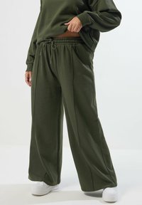 Olivgrön vida byxor med elastisk midja och dragsko, tillverkade av ett mjukt tyg, kombinerade med vita sneakers.