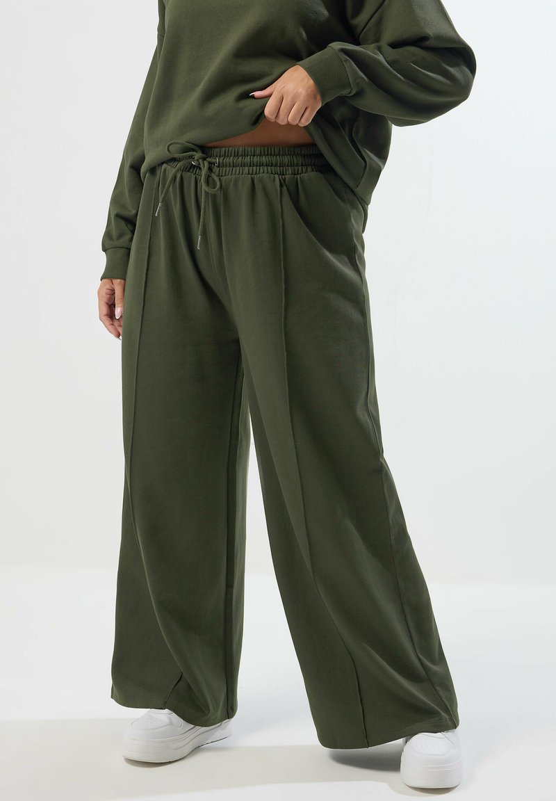 Olivgrön vida byxor med elastisk midja och dragsko, tillverkade av ett mjukt tyg, kombinerade med vita sneakers.