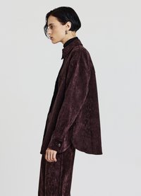 Camicia cremisi realizzata in tessuto morbido e strutturato, con collo a camicia, chiusura con bottoni e spacchi laterali per una vestibilità comoda.