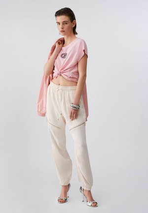 Donna con top corto rosa legato, pantaloni jogger color crema, indossa tacchi alti argentati e braccialetti, tiene una giacca rosa abbinata appoggiata sulla spalla.