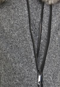Chaqueta gris de felpa con cierre de cremallera, fabricada con un material texturizado, que cuenta con una cremallera negra y un tirador metálico. Detalle de capucha esponjosa en el cuello.