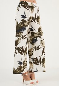 Bredere broek in een lichte stof, met een witte achtergrond en grote groene en marineblauwe bladdessins. Elastische tailleband voor comfort.