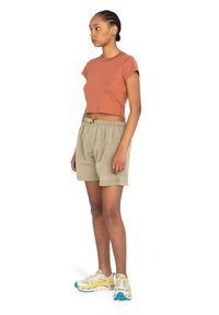 Hnedo-červený crop top s krátkymi rukávmi, skombinovaný s béžovými kockovanými šortkami s elastickým pásom a žltou šnúrkou. Svetlé tenisky.