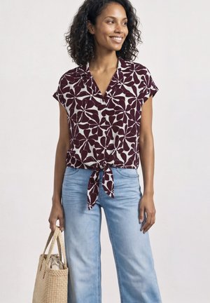 Uśmiechnięta kobieta w krótkim rękawie, zapinanej na guziki koszuli w kwiatowy wzór, związanej w talii, w jasnoniebieskich dżinsach, trzymająca plecioną torbę.