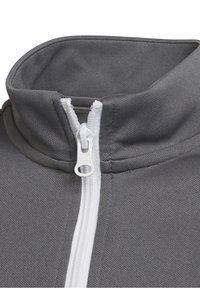 Grå zip-jakke med høj krave, der har en hvid lynlås. Stoffet har en struktureret finish, og stingdetaljer er synlige.