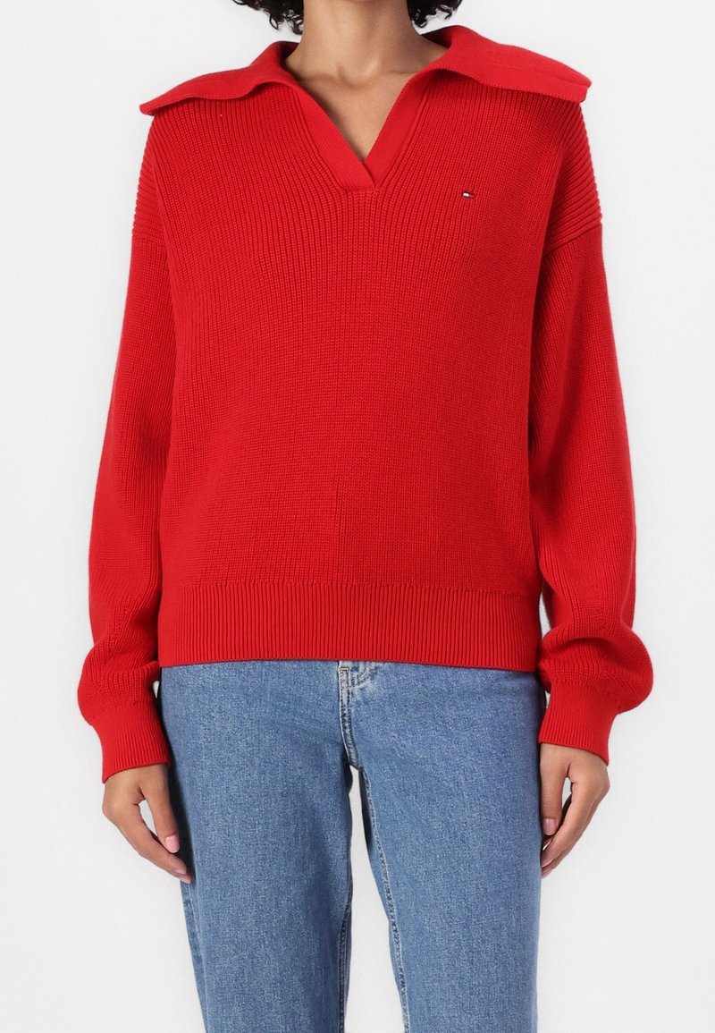 Red knit sweater with a deep V-neck and large collar, featuring ribbed cuffs and hem, paired with light blue jeans.