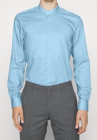 Camicia azzurro chiaro a maniche lunghe con colletto alla mandarino, realizzata in tessuto morbido. Abbinata a pantaloni grigi con motivo. Bottoni visibili davanti.