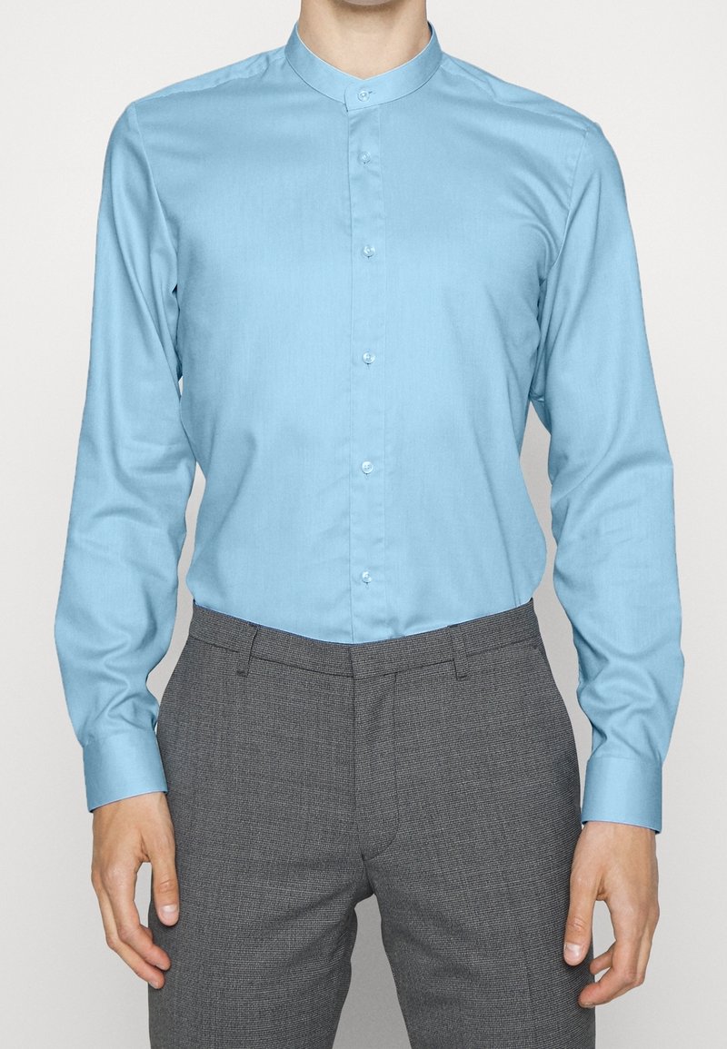 Camicia azzurro chiaro a maniche lunghe con colletto alla mandarino, realizzata in tessuto morbido. Abbinata a pantaloni grigi con motivo. Bottoni visibili davanti.