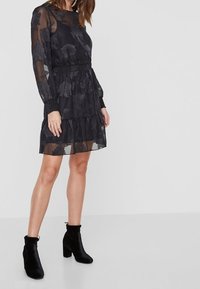 Svart klänning med blommönster, långa ärmar, figursydd midja och lager-på-lager kjol. Genomskinligt tyg med texturerade detaljer på ärmarna. Svarta klackstövletter.