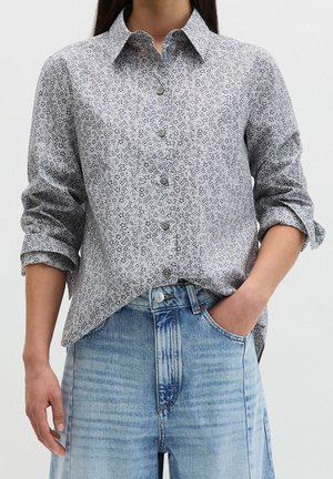 Kvinde iført en langærmet, knappet lysgrå blomsterbluse med opkrogede ærmer og lyseblå højtaljede jeans, med den ene hånd i lommen.