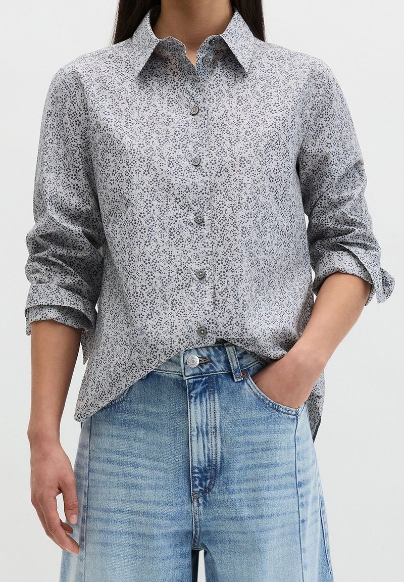 Femme portant une chemise fleurie gris clair à manches longues boutonnée, manches retroussées, et un jean bleu clair taille haute, une main dans la poche.