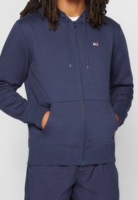 Felpa con zip di colore navy realizzata in tessuto morbido; presenta tasche frontali, cappuccio con lacci e piccolo logo sul petto; include polsini e bordo a costine.