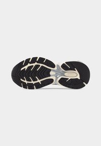 Semelle d'une chaussure de sport avec une bande de roulement en caoutchouc noir et beige, comprenant un élément de soutien médio-pied argenté et un motif de grip texturé.