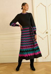 Rochie neagră cu mâneci lungi, având un model colorat în zigzag în nuanțe de roz și teal. Curea la talie, decolteu cu volane și țesătură texturată.