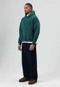 Sweat-shirt à capuche vert avec une poche frontale, associé à un jean bleu foncé ample et des bottes de randonnée marron avec des accents rouges.
