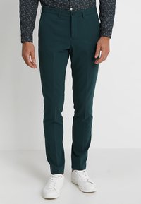 Teal uske hlače slim fit od glatkog materijala, s prednjim zatvaračem i kopčanjem na gumb. Nosene su s košuljom s uzorkom i bijelim tenisicama.