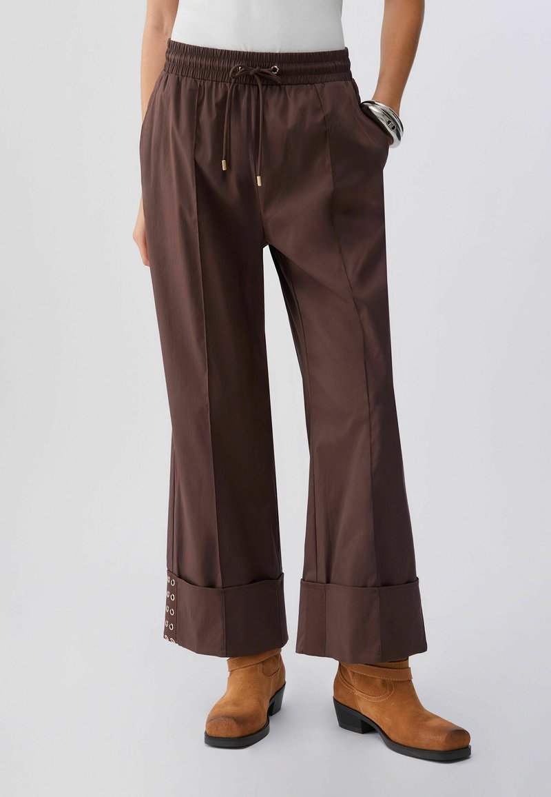 Pantalon marron à jambes larges avec ourlets retroussés et détails à œillets lacés, porté avec des bottines en daim camel et un haut blanc.
