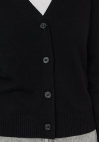 Czarny dziergany kardigan z dekoltem w serek, sześcioma guzikami, ściągaczami na mankietach i dolnym brzegu. Materiał ma delikatną fakturę. Nosi się go na jasną koszulę.