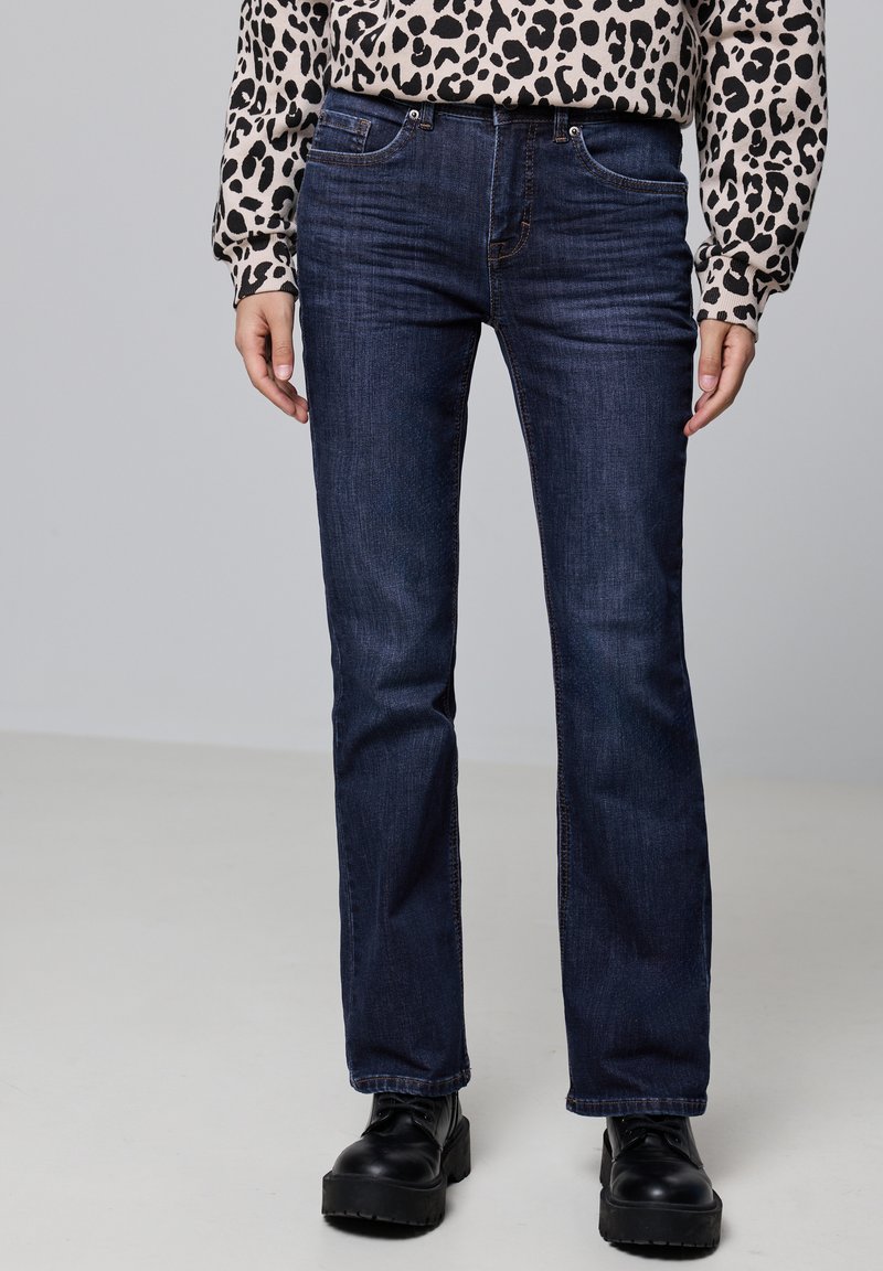 Jeans de mezclilla azul oscuro con corte de pierna recta, diseño estándar de cinco bolsillos y costuras resistentes. Combinados con una camisa de manga larga con estampado.