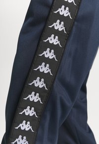 Marina atletiska byxor har en sidopanel med en upprepande vit logotypdesign på en svart bakgrund, vilket visar en slät tygtextur.