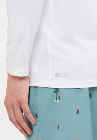Hvit langermet skjorte med UPF 50+ merket nederst, sammen med blå shorts med palme- og strandmønster.