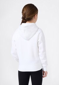 Joven con cabello trenzado marrón, vistiendo una sudadera blanca con capucha y pantalones negros, de espaldas contra un fondo blanco liso.