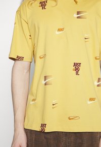 Żółta bawełniana koszulka z powtarzającymi się logo Nike i napisem "JUST DO IT." w bordowym kolorze, krótkie rękawy, standardowy krój.