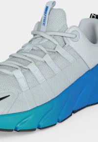 Chaussure de sport en mesh blanc avec des brides gris clair, semelle dégradée bleue, et "NIKE FREE" imprimé en bleu sur la languette.