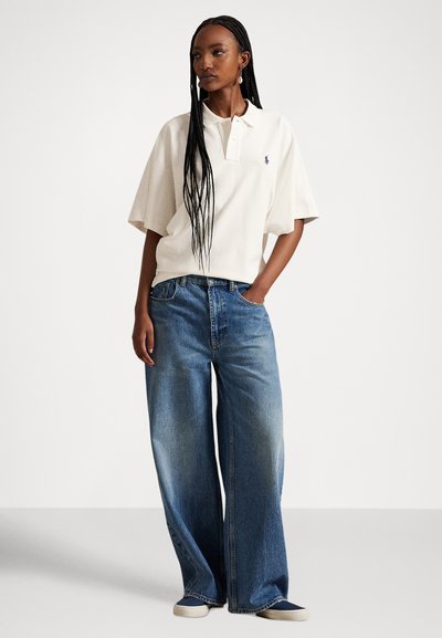 Balti trumpomis rankovėmis polo marškinėliai su apykakle ir logotipu, derinami su plačiomis kojų mėlynos spalvos džinsais. Lygus audinys ir laisvas įpjovimas.