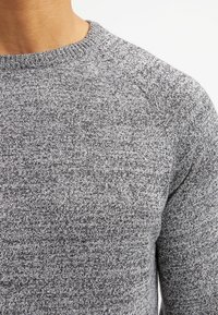 Szary sweter z dzianiny o fakturze z plamkowanym wzorem. Dekolt ma standardowy fason okrągły, a materiał wydaje się być miękki i elastyczny.