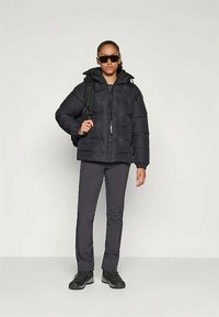 Άτομο φορώντας μαύρη πούφφα τζάκετ, σκούρα παντελόνια, μπότες πεζοπορίας, γυαλιά ηλίου και σακίδιο, σε στάση ενάντια σε απλό φωτεινό φόντο.