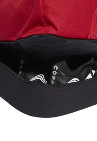 Bolso rojo con un compartimento negro con cremallera que contiene calzado negro. El calzado presenta un logotipo y acentos de diseño en blanco.