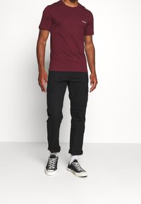 T-shirt corto color bordeaux con colletto tondo e logo bianco sul petto, abbinato a pantaloni cargo neri con tasche laterali e sneakers nere.