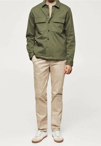 Oliivinvihreä napillinen takki, jossa kaksi rintataskua, beiget slim fit -housut ja valkoiset lenkkarit ruskeilla yksityiskohdilla. Sileä kangaspinta.