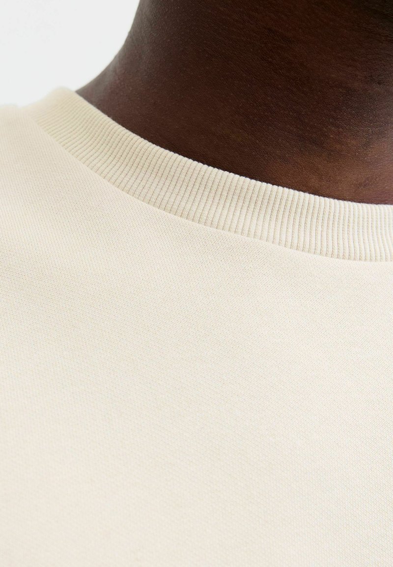 Pull en beige texturé avec un col ras du cou côtelé, gros plan sur la zone de l'épaule mettant en évidence le tissu lisse et uniforme.
