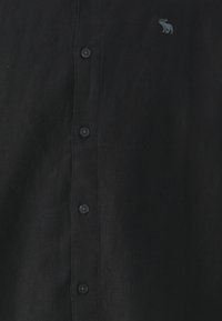 Camisa de lino negra con un cierre frontal de botones, seis botones negros y un pequeño logo de alce bordado en el pecho.