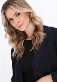 Cardigan tessuto blu navy con apertura frontale, indossato sopra una camicia nera. Il modello ha capelli mossi e un trucco sobrio, rivolto leggermente verso la fotocamera.
