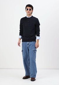 Felpa nera con logo in rilievo "TOMMY HILFIGER", camicia con colletto azzurra sotto, jeans cargo blu e scarpe marroni.