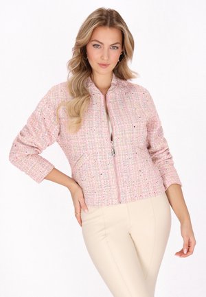 Frau mit langen blonden Haaren, die eine rosa strukturierte Jacke mit Reißverschluss und cremefarbene Hose trägt, posiert mit einer Hand auf der Hüfte.