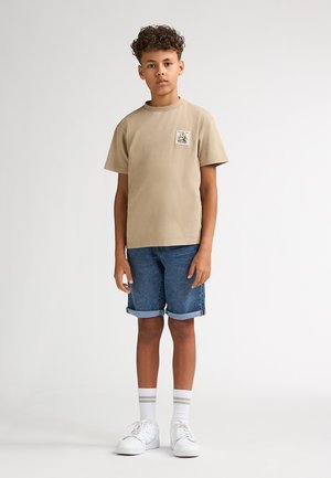 Beige short-sleeve t-shirt with a chest pocket patch, paired with rolled denim shorts, white sneakers, and striped white socks.