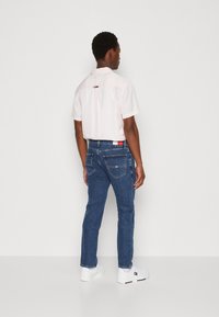 Mann som står med ryggen til, iført en lys rosa kortermet skjorte, blå jeans med logo-patch, og hvite joggesko mot en ensfarget vegg.