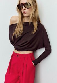 Top marrone a maniche lunghe con scollo a barca abbinato a pantaloni rosa plissettati a vita alta con cintura. La modella indossa occhiali da sole oversize con montatura tartarugata.